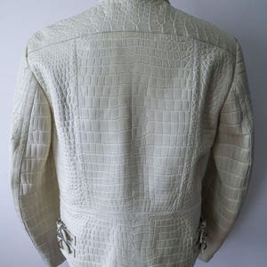 May include: Cream-colored leather jacket with a crocodile skin texture. The jacket has a stand-up collar, a zipper closure, and decorative metal accents near the waist. The back of the jacket is visible, showcasing the detailed embossed pattern.