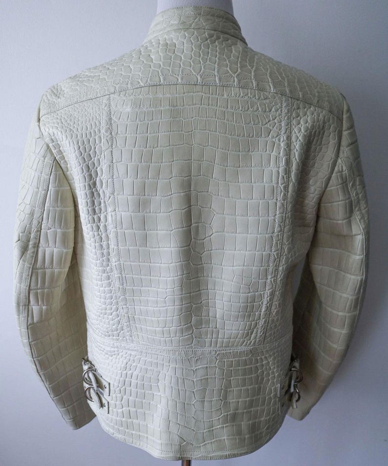 May include: Cream-colored leather jacket with a crocodile skin texture. The jacket has a stand-up collar, a zipper closure, and decorative metal accents near the waist. The back of the jacket is visible, showcasing the detailed embossed pattern.