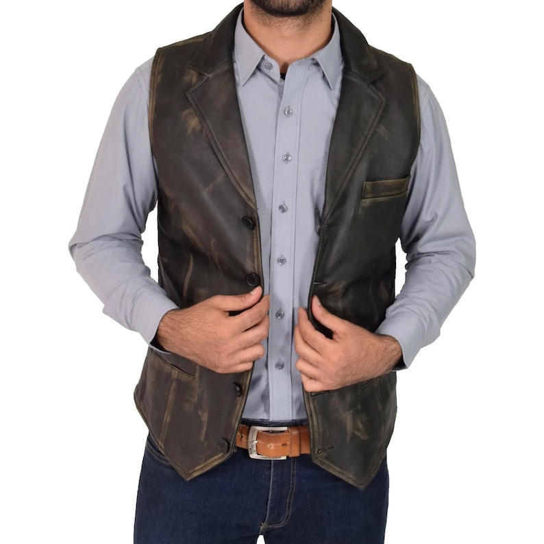 May include: A dark brown leather vest with a notched lapel and three buttons. The vest has a distressed finish and a small pocket on the left side. The person is wearing a light blue button-down shirt and blue jeans.