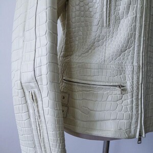 May include: Cream-colored leather jacket with a textured, crocodile-like pattern. Features a zipper closure, multiple pockets, and a tailored fit. The jacket is displayed on a mannequin, showcasing its design and details.