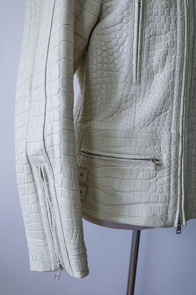 May include: Cream-colored leather jacket with a textured, crocodile-like pattern. Features a zipper closure, multiple pockets, and a tailored fit. The jacket is displayed on a mannequin, showcasing its design and details.