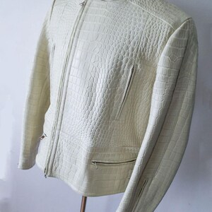 May include: Cream-colored leather jacket with a textured, crocodile-like pattern. The jacket features a zipper closure, multiple pockets, and zippers on the sleeves. The collar is a stand-up style.