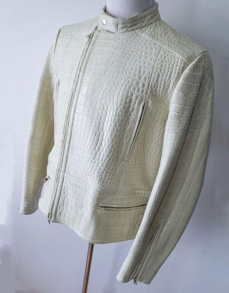 May include: Cream-colored leather jacket with a textured, crocodile-like pattern. The jacket features a zipper closure, multiple pockets, and zippers on the sleeves. The collar is a stand-up style.