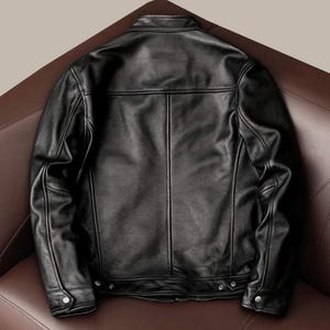 May include: A black leather jacket with a classic design. The jacket features a stand-up collar, a front zipper closure, and multiple pockets. The back of the jacket is visible, showcasing the tailored fit and stitching details. The jacket is displayed on a brown leather surface.