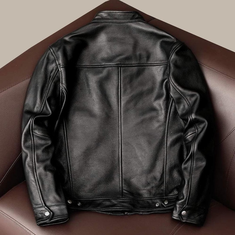 May include: A black leather jacket with a classic design. The jacket features a stand-up collar, a front zipper closure, and multiple pockets. The back of the jacket is visible, showcasing the tailored fit and stitching details. The jacket is displayed on a brown leather surface.