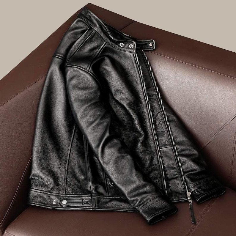 May include: A black leather jacket with a zipper closure, snap buttons, and a classic collar. The jacket is displayed on a brown leather surface, showcasing its sleek design and texture. The jacket's details include a zippered front and a tailored fit.