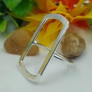 May include: A silver ring with a rectangular open design. The ring is set against a white surface with small brown stones and colourful flowers in the background. The ring's design is modern and minimalist.