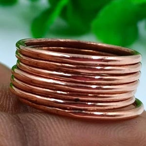 May include: A collection of copper-coloured rings. The rings are stacked, creating a layered effect. The rings have a warm, reddish-brown hue. The background features green foliage and a white surface.