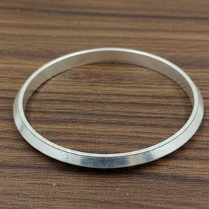 Könnte beinhalten: Ein silberfarbenes Armreif-Armband mit einer flachen, polierten Oberfläche. Das kreisförmige Armband liegt auf einer braunen Holzoberfläche. Das Armreif ist ein schlichtes, elegantes Schmuckstück.