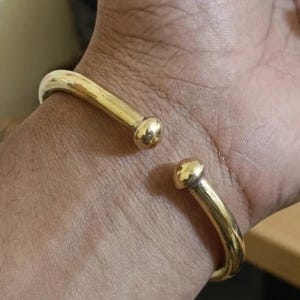 以下が含まれることがあります： 滑らかで光沢のある仕上げが施されたゴールドトーンの金属製カフブレスレット。円形で開口部のあるデザインが特徴です。ブレスレットの両端は丸みを帯びた球状の留め具になっており、それらが向かい合うことで、連続した曲線を描くバンド構造を作り出しています。