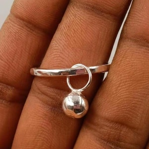 Puede incluir: Un anillo de plata con un pequeño dije de bola colgante. El anillo tiene un diseño sencillo, con una banda delgada y un pequeño dije redondo que cuelga libremente. El anillo se muestra en un dedo.