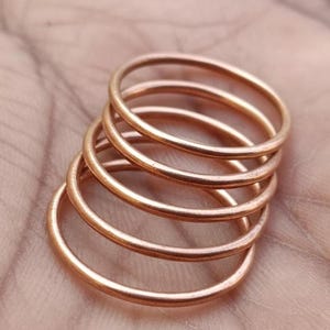Può includere: Una pila di cinque anelli sottili color rame. Gli anelli sono circolari e sembrano essere di metallo. Gli anelli sono impilati l'uno sull'altro, creando un effetto a strati. Gli anelli poggiano su una superficie marrone chiaro.