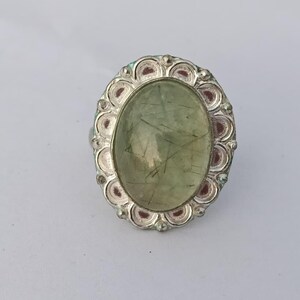 May include: A silver ring featuring a large, oval-shaped, pale green cabochon gemstone with visible dark needle-like inclusions. The stone is set within a decorative, scalloped silver frame that has a series of small, circular indentations.