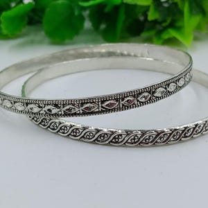 K&ouml;nnte beinhalten: Zwei silberne Armreifen mit aufwendigen Designs. Einer der Armreifen hat ein sich wiederholendes Muster aus blattartigen Formen, der andere hat rautenf&ouml;rmige Akzente. Die Armreifen werden auf einer wei&szlig;en Oberfl&auml;che mit gr&uuml;nem Laub im Hintergrund gezeigt.