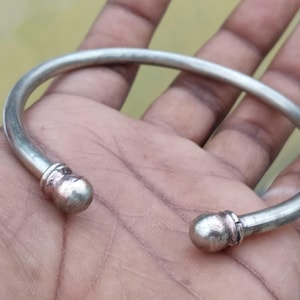 May include: A silver-coloured, open cuff bracelet with rounded ball ends. The bracelet is held in a hand, showcasing its simple design. The metal has a slightly aged appearance.