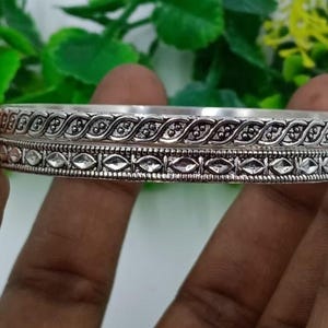 K&ouml;nnte beinhalten: Ein silberfarbenes Armreif-Armband mit aufwendigen, detaillierten Mustern. Das Design umfasst eine Reihe von verbundenen blattartigen Formen und geometrischen Mustern. Das Armband wird in einer Hand gehalten und zeigt seine kunstvolle Handwerkskunst.