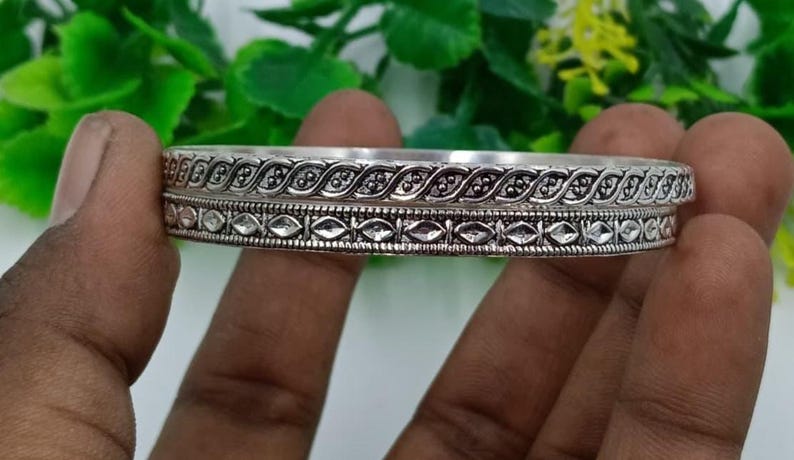 K&ouml;nnte beinhalten: Ein silberfarbenes Armreif-Armband mit aufwendigen, detaillierten Mustern. Das Design umfasst eine Reihe von verbundenen blattartigen Formen und geometrischen Mustern. Das Armband wird in einer Hand gehalten und zeigt seine kunstvolle Handwerkskunst.