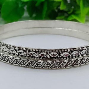 K&ouml;nnte beinhalten: Zwei silberne Armreifen mit detaillierten Mustern. Die Armreifen haben eine polierte Oberfl&auml;che und sind mit einem sich wiederholenden Design aus Diamanten und Bl&auml;ttern verziert. Der Schmuck wird auf einer wei&szlig;en Oberfl&auml;che pr&auml;sentiert.