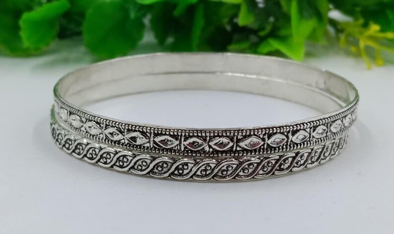 K&ouml;nnte beinhalten: Zwei silberne Armreifen mit detaillierten Mustern. Die Armreifen haben eine polierte Oberfl&auml;che und sind mit einem sich wiederholenden Design aus Diamanten und Bl&auml;ttern verziert. Der Schmuck wird auf einer wei&szlig;en Oberfl&auml;che pr&auml;sentiert.
