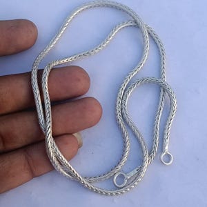 Pode incluir: Um colar de corrente de cor prateada com um padrão de elos trançados. A corrente é flexível e inclui dois pequenos elos circulares nas extremidades para fecho. A superfície metálica tem um acabamento brilhante e refletor em todo o design.