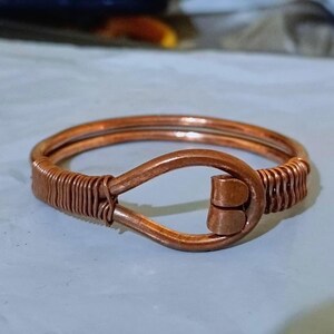 May include: A copper wire bracelet featuring a double-strand design with a central teardrop-shaped loop closure. The ends of the wire are tightly coiled and wrapped around the main band, creating a secure and structured metallic accessory.