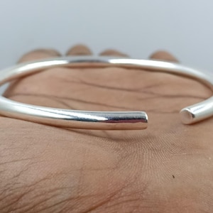 Può includere: Un braccialetto a bracciale argentato, aperto. Il braccialetto ha un design liscio e arrotondato e misura circa 6,35 cm di diametro. Le estremità del braccialetto sono arrotondate e lucidate. Il braccialetto è mostrato su uno sfondo color carne.
