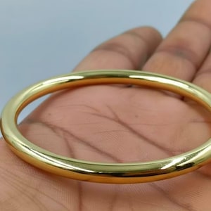 May include: A gold-colored bangle bracelet. The circular bracelet is smooth and shiny, reflecting light. The jewelry is held in a hand, with the background out of focus.