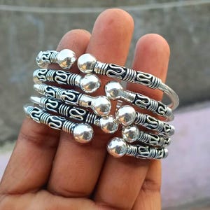 Può includere: Una pila di cinque bracciali aperti in metallo color argento con terminali a sfera arrotondati. Ogni bracciale presenta un motivo ripetitivo di solchi ossidati scuri e segmenti in rilievo color argento lungo la fascia.