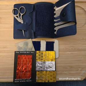 May include: A dark blue folding sewing kit case containing gold-toned scissors, a spool of dark blue thread, and a needle book labeled This STITCH KIT has NEEDLES FOR YOUR MANY SEWING NEEDS with gold and red foil accents and assorted needles.