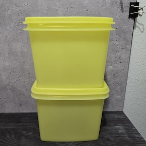 May include: Two yellow, square-shaped food storage containers with matching lids are stacked on top of each other. The containers are made of a translucent plastic material. The lids fit snugly on top of the containers.
