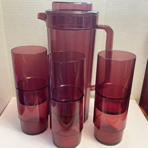 May include: A translucent deep purple plastic pitcher with a matching lid and handle, accompanied by three tall, cylindrical drinking glasses in the same matching deep purple hue, each featuring a distinct horizontal tiered design.