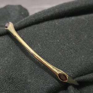 May include: A gold-toned metal bangle bracelet featuring a smooth, tapered design with two small, dark red oval-shaped stones set at each end. The bracelet has a polished finish and is displayed against a dark, textured fabric background.