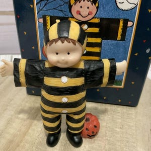 May include: A figurine of a child wearing a black and yellow striped bumblebee costume with a matching hood, three white buttons down the front, and black shoes. A small orange jack-o'-lantern sits at the feet of the figurine, which stands in front of a box.