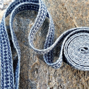 Peut inclure: Une sangle tissée à motifs bleus et blancs, enroulée et posée sur une surface de pierre texturée. La sangle est faite d'un matériau doux et présente un motif détaillé et répétitif. La partie enroulée forme une spirale.