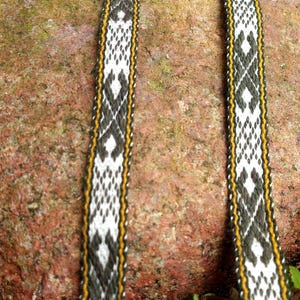 May include: Close-up of two woven straps with a geometric pattern in black, white, and yellow. The straps are lying on a reddish-brown rock, with green foliage visible in the background. The straps have a repeating diamond and cross design.
