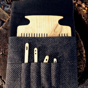 May include: A dark grey woven fabric case with a wooden button closure, containing wooden weaving tools. The tools include a large comb, a small shuttle, and several needle-like tools. The case is open, displaying the tools.