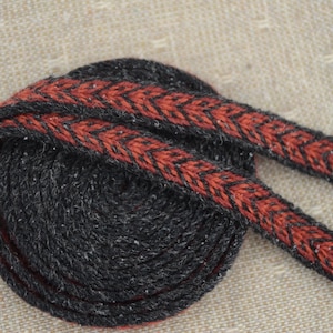 Puede incluir: Un ribete tejido negro y gris texturizado enrollado, con un patrón de chevrón rojo y negro. El ribete probablemente está hecho de hilo o tela y es adecuado para coser, hacer manualidades o embellecer varios proyectos.