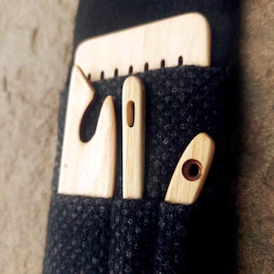 May include: Four wooden weaving tools, including a comb, a needle, and two shuttles, are displayed in a dark grey fabric pocket. The tools are made of light-coloured wood. The image is a close-up shot, showing the tools in detail.
