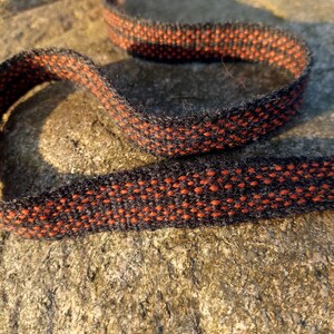 May include: Close-up of a woven ribbon with a repeating pattern of orange dots on a black background. The ribbon is curved and lies on a rough, textured stone surface. The ribbon's edges are neatly finished.