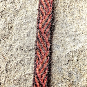 May include: Close-up of a woven textile strip with a chevron pattern. The strip features alternating bands of red, orange, and dark brown. The background is a textured, light brown stone surface.