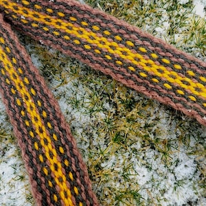 May include: Close-up of a woven textile with a repeating pattern of brown, yellow, and dark brown. The textile is lying on a bed of green grass and snow. The woven material appears to be a belt or strap.