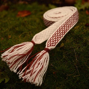 May include: A woven belt with a cream-colored base and a burgundy geometric pattern. The belt has tassels at each end, with cream and burgundy threads. The belt is displayed on a bed of green moss.