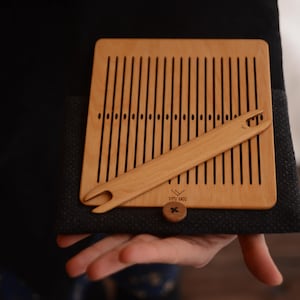 May include: A wooden weaving loom and shuttle set. The loom is rectangular with vertical slots, and the shuttle is a long, thin piece with a hook. The set is light brown and rests on a dark fabric.