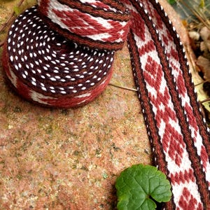 Puede incluir: Un cinturón textil tejido en tonos burdeos, blanco y marrón, con un patrón geométrico de diamantes. El cinturón está enrollado y parcialmente deshecho, mostrando su intrincado diseño. La imagen está ambientada sobre un fondo natural con follaje verde.