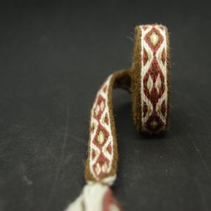 Può includere: Un tessuto marrone tessuto con un motivo geometrico in rosso, crema e oro. Il tessuto è curvo, con un anello a un'estremità e una lunga e stretta striscia che si estende da esso. Lo sfondo è scuro.