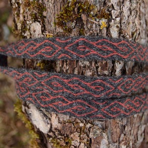 Può includere: Fascia per capelli tessuta con un motivo geometrico grigio e rosso. La fascia è avvolta attorno a un tronco d'albero, mostrando l'intricato design e il contrasto cromatico. Lo sfondo è sfocato, evidenziando la fascia.