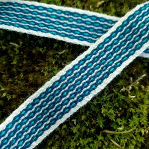 Op de afbeelding: Close-up van twee geweven linten met een witte basis en een herhalend patroon van turquoise en marineblauwe ruiten. De linten liggen op een bed van groen mos, waardoor een natuurlijke en gestructureerde achtergrond ontstaat. De linten worden waarschijnlijk gebruikt voor knutselen of decoratie.