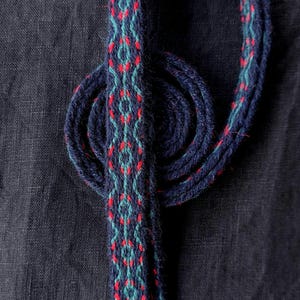 May include: A woven textile with a repeating pattern of red and teal diamonds on a navy blue background. The textile is looped and coiled, showcasing the intricate design. The background is a dark navy blue fabric.