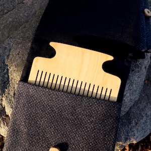 May include: A wooden comb with a unique shape is nestled in a dark gray fabric case. The comb is light-colored wood with evenly spaced teeth. The case has a button closure and a textured appearance, suggesting a natural fiber.