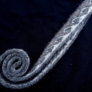 May include: A coiled, woven textile with a geometric pattern in gray, white, and brown. The textile is on a dark background, with the coiled end in the lower left corner. The pattern is a repeating diamond design.